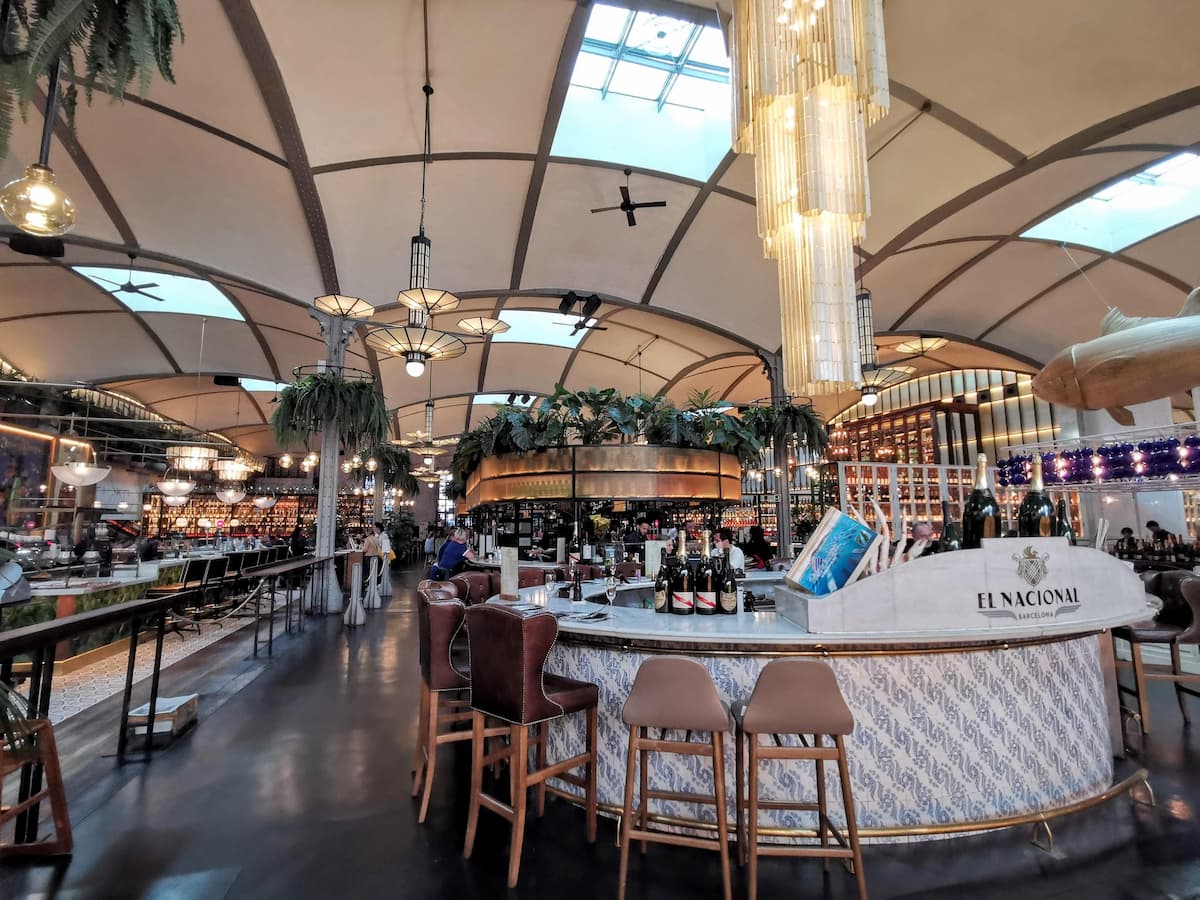 Interior of El Nacional Barcelona showing the grand central bar with vaulted skylights, hanging chandeliers and tropical plants